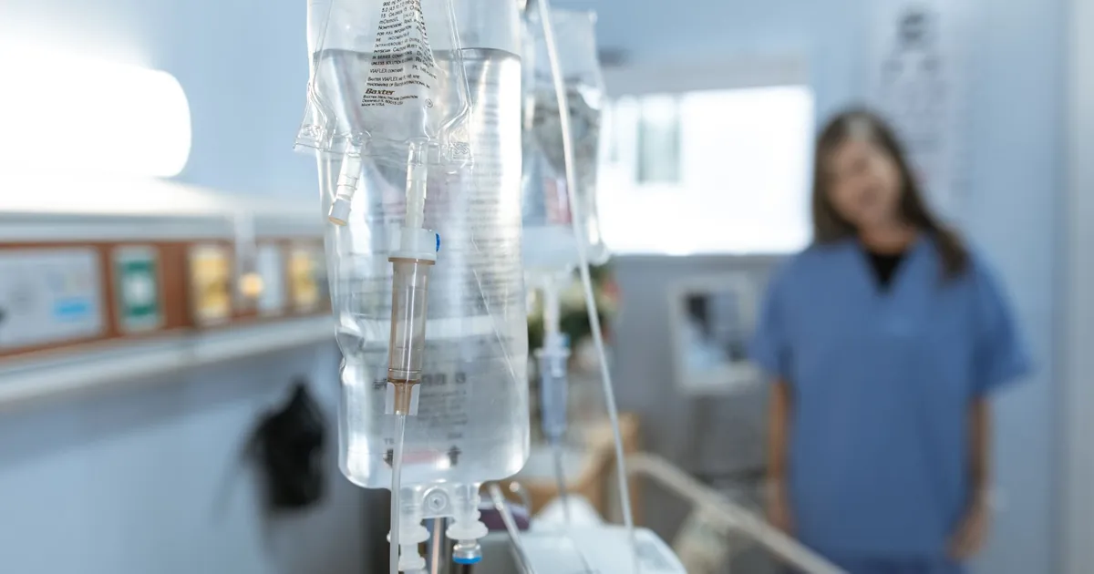 An IV drip in a hospital room, with a nurse out of focus behind it