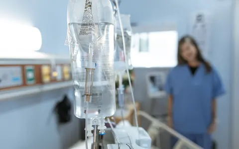 An IV drip in a hospital room, with a nurse out of focus behind it
