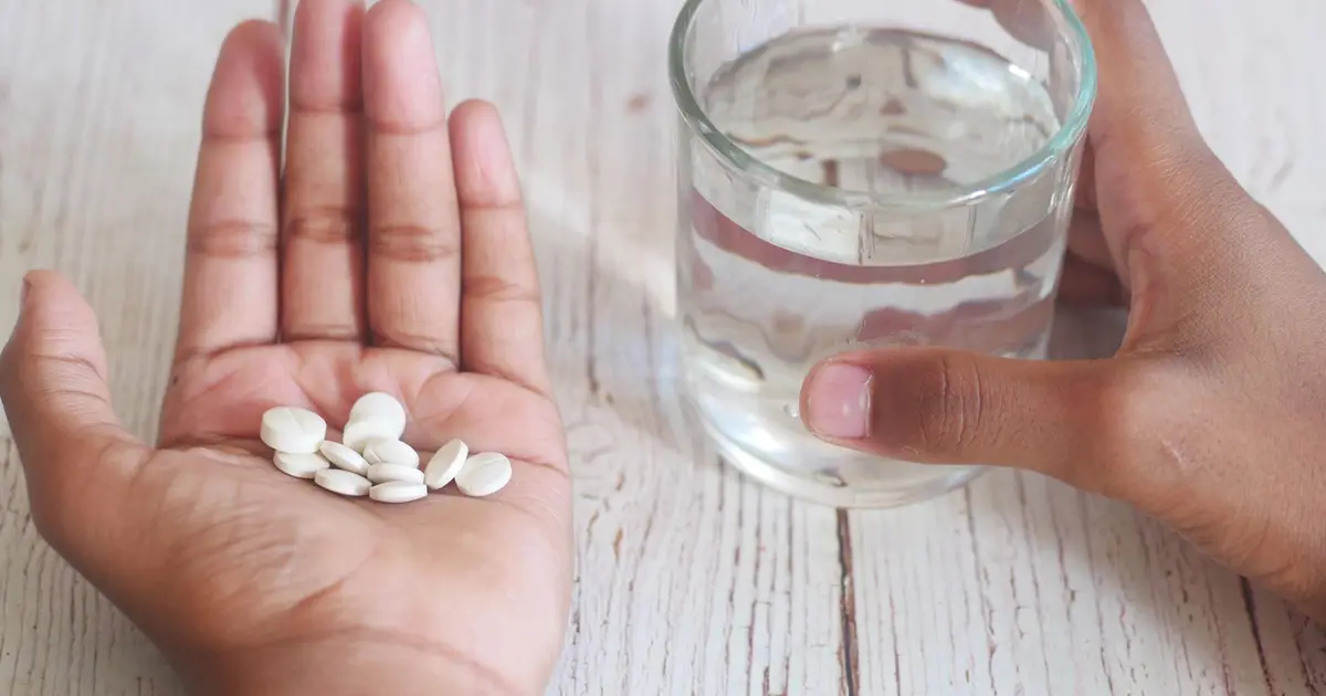 A close-up of hands holding medication tablets and a glass of water on a wooden surface