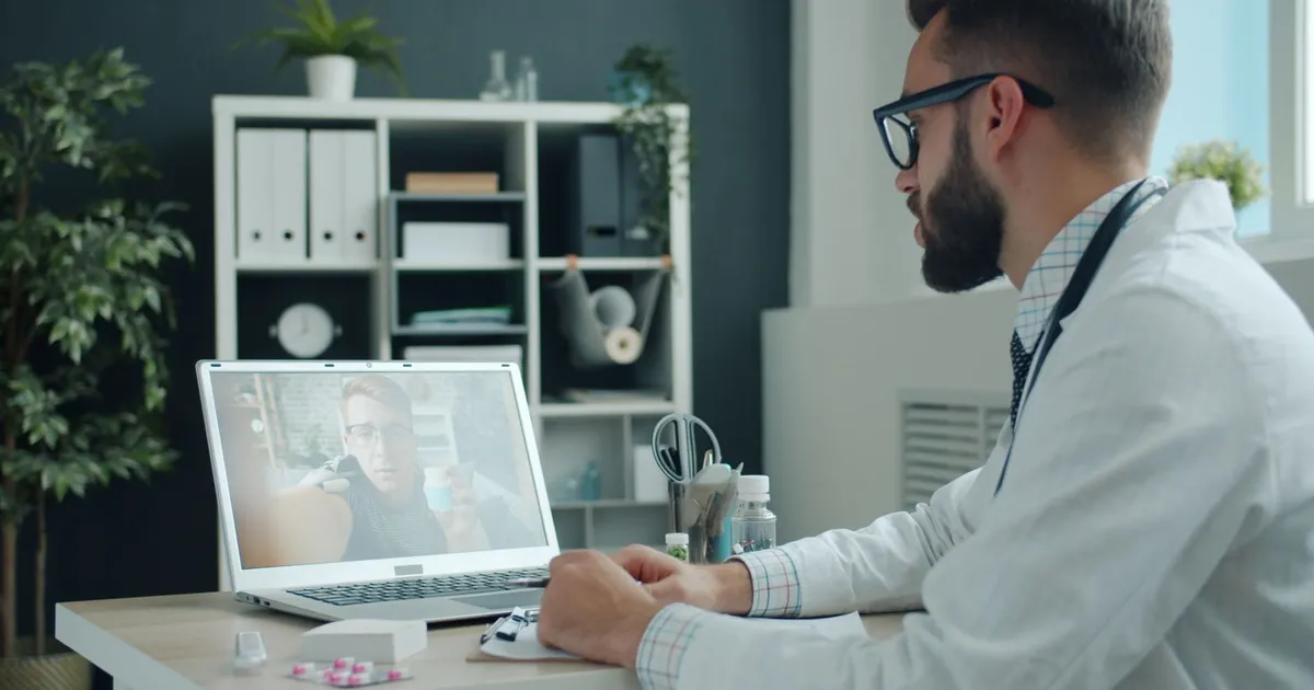 A doctor in a white coat on a video call with a patient on a laptop