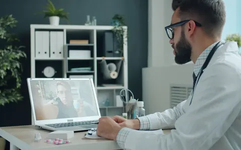 A doctor in a white coat on a video call with a patient on a laptop