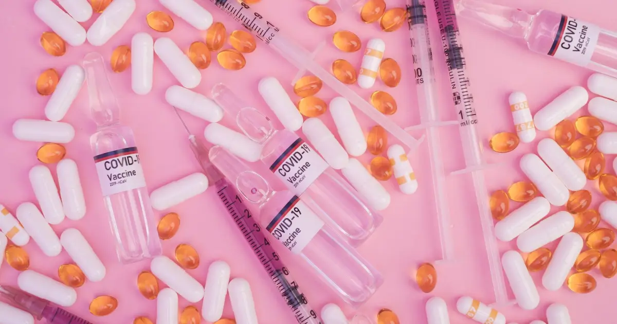 Top view of various medicine capsules scattered on pink table with syringes and ampoules of COVID 19 vaccine