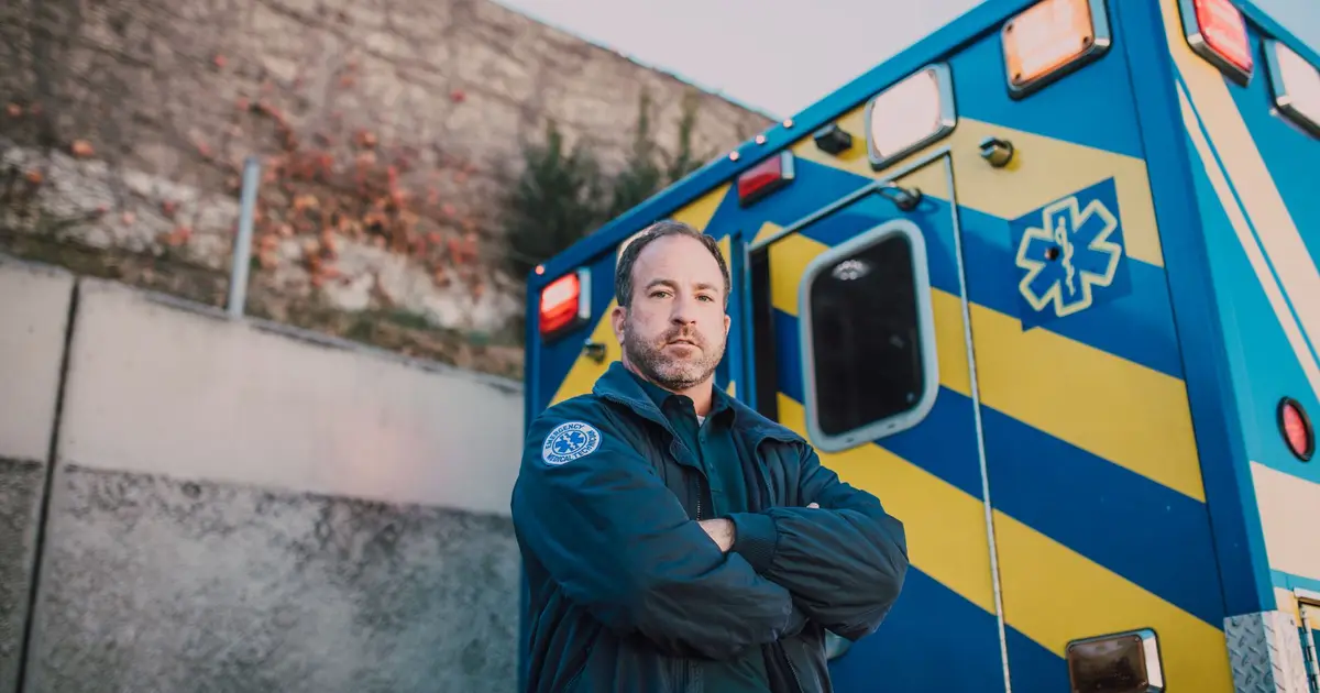 Male paramedic standing confidently beside a blue and yellow ambulance outdoors