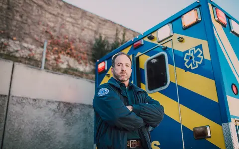 Male paramedic standing confidently beside a blue and yellow ambulance outdoors
