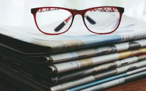 Red eyeglasses resting on a stack of newspapers symbolize reading and information