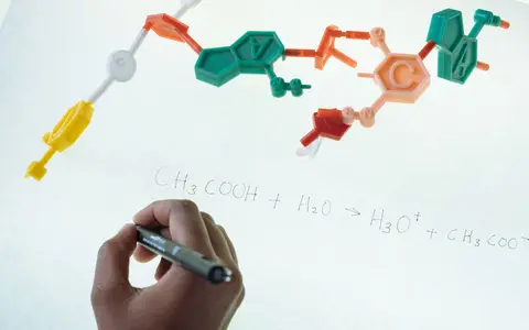 Anonymous chemist writing with marker on illuminated magnetic board formula of Hydronium and Acetate production after studying plastic model of molecule in laboratory